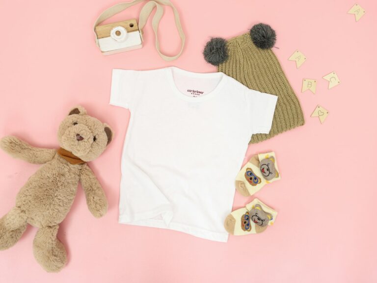 Soft white baby clothes laid flat on a pink surface next to a teddy bear and a bear hat